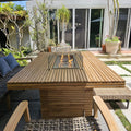 Louvre teak fire pit dining table with a lit burner and glass wind guard, surrounded by woven chairs on a garden patio.