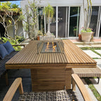 Louvre teak fire pit dining table with a lit burner and glass wind guard, surrounded by woven chairs on a garden patio.