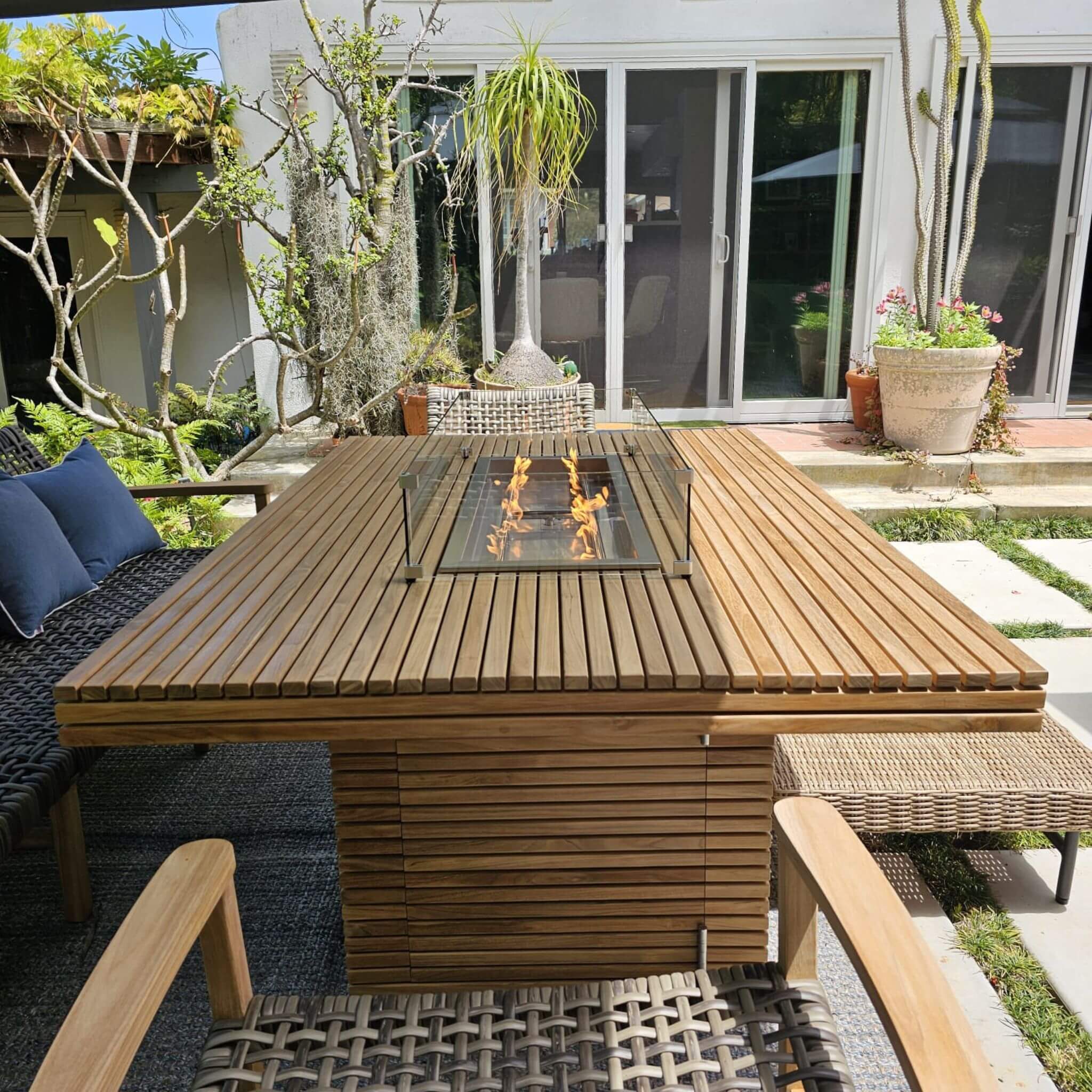 Louvre teak fire pit dining table with a lit burner and glass wind guard, surrounded by woven chairs on a garden patio.