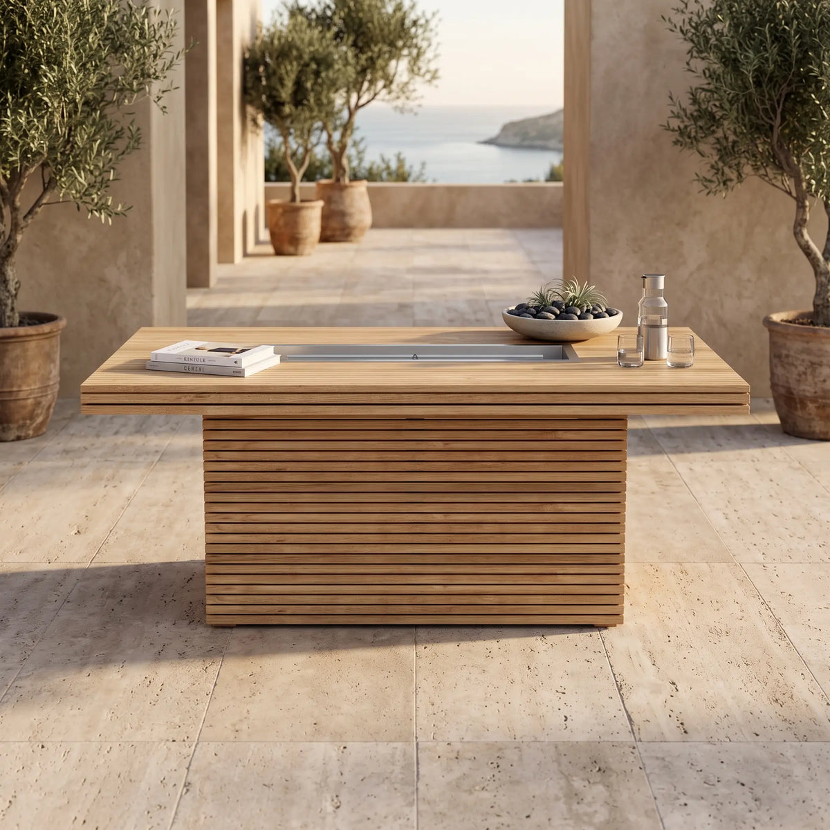 Front view of a teak fire table with an unlit central burner, books and a tray on a stone terrace.