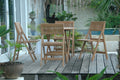 Round folding teak bistro table with four folding chairs on a wooden deck beside a pond.