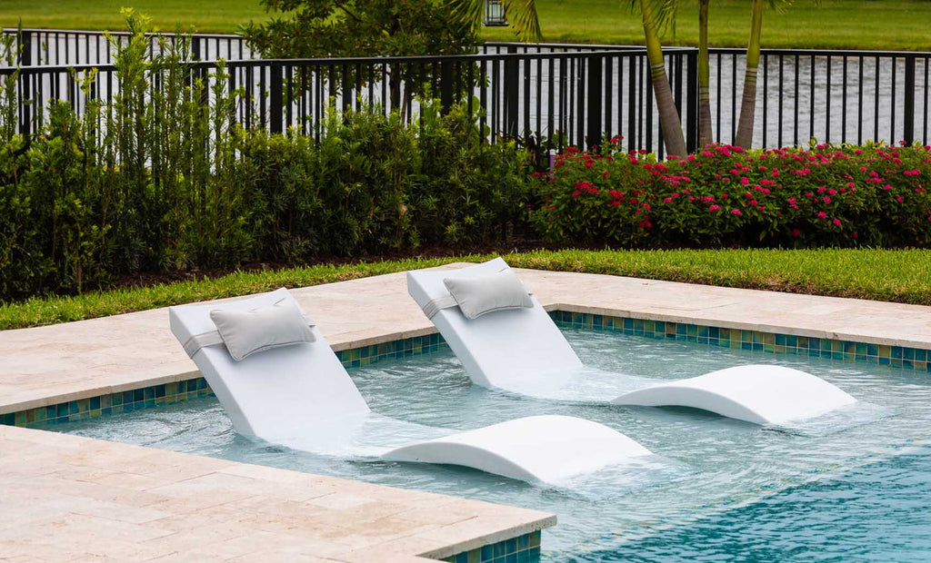 Two White Signature Chaises with headrest pillows on a shallow pool ledge; lifestyle styling example.