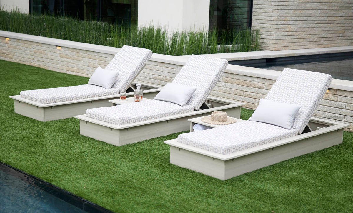 Three Ledge Lounger Echo chaises with pale woodgrain-look frames and geometric-pattern cushions arranged on a lawn beside a pool.