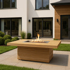 Teak fire pit table with a lit rectangular burner on a patio beside a modern house and lawn.