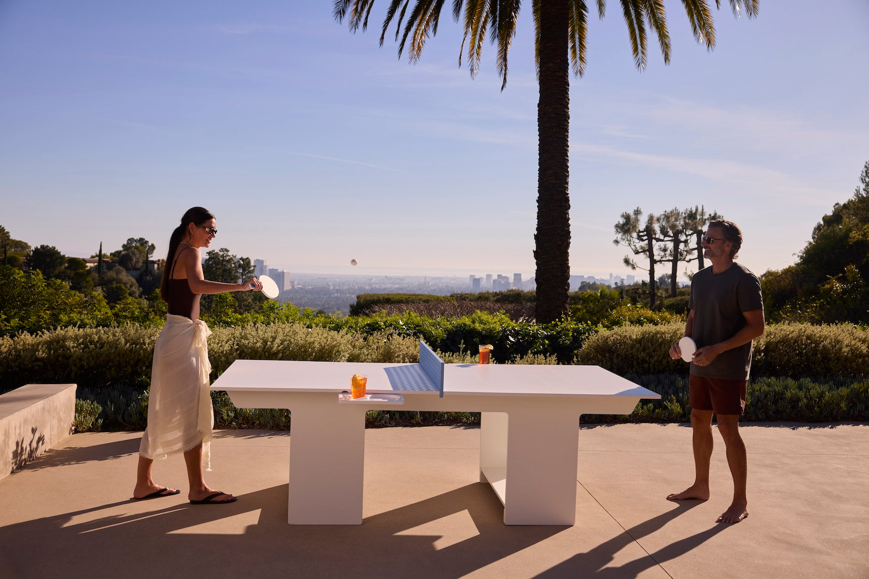 Two people playing at the Ledge Lounger Table Tennis Set outdoors with drinks on the table, surrounded by greenery