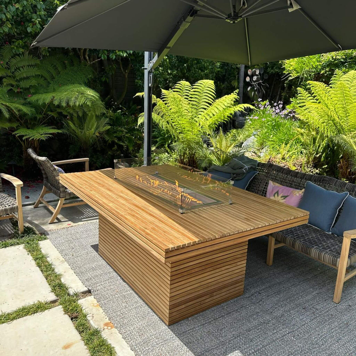 Angled garden view of the Louvre fire table beside woven seating with blue cushions.