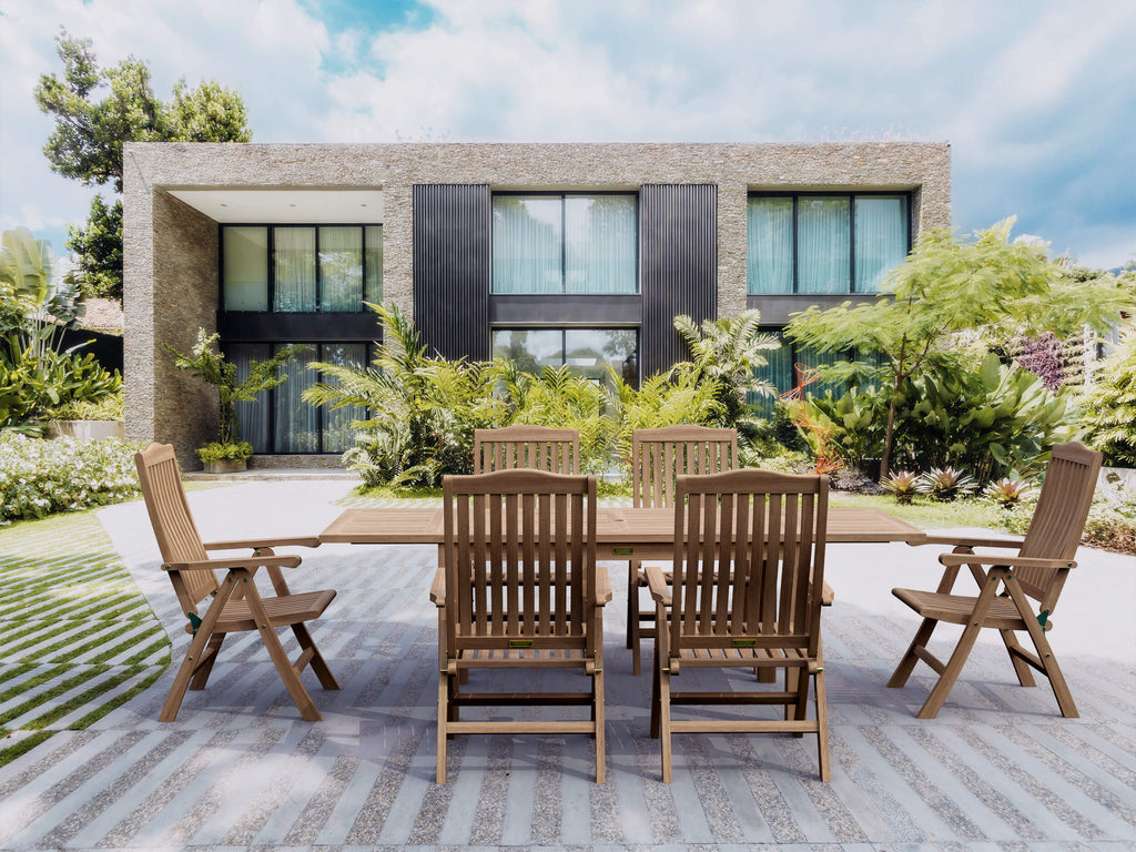 Bahama Katana teak extension dining table with six reclining teak armchairs on a paved terrace in front of a modern stone-and-glass house surrounded by tropical planting