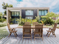 Bahama Katana teak extension dining table with six reclining teak armchairs on a paved terrace in front of a modern stone-and-glass house surrounded by tropical planting