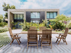 Bahama Katana teak extension dining table with six reclining teak armchairs on a paved terrace in front of a modern stone-and-glass house surrounded by tropical planting