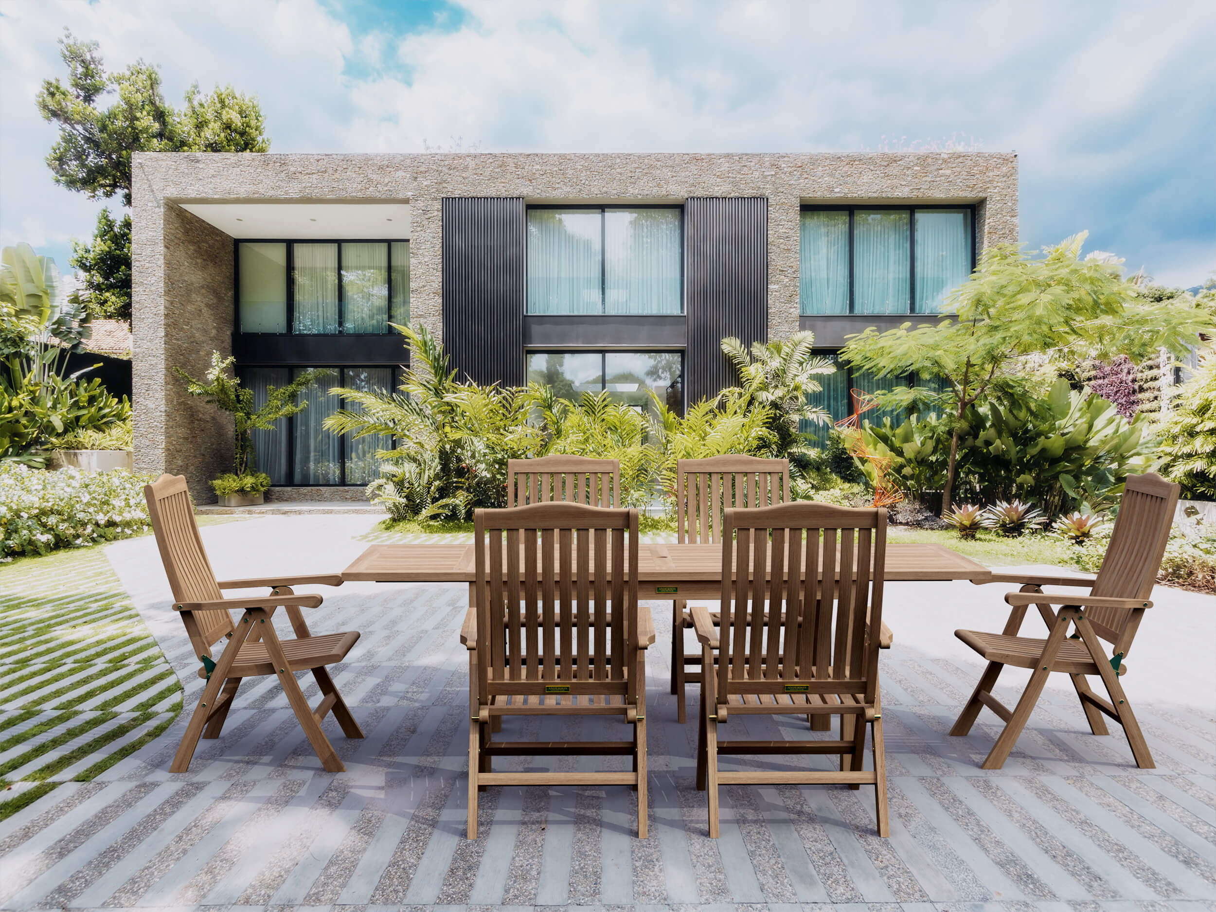 Bahama Katana teak extension dining table with six reclining teak armchairs on a paved terrace in front of a modern stone-and-glass house surrounded by tropical planting