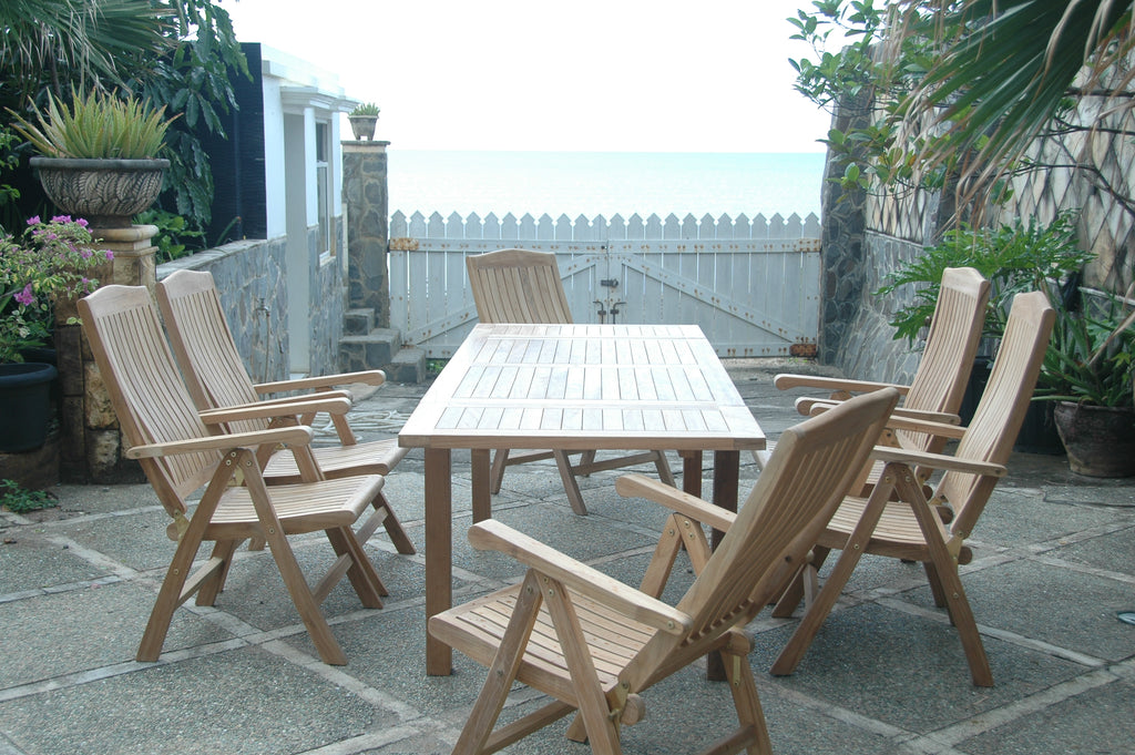Bahama Katana teak extension dining table and six reclining armchairs on a seaside stone patio, with a white picket gate and the ocean behind