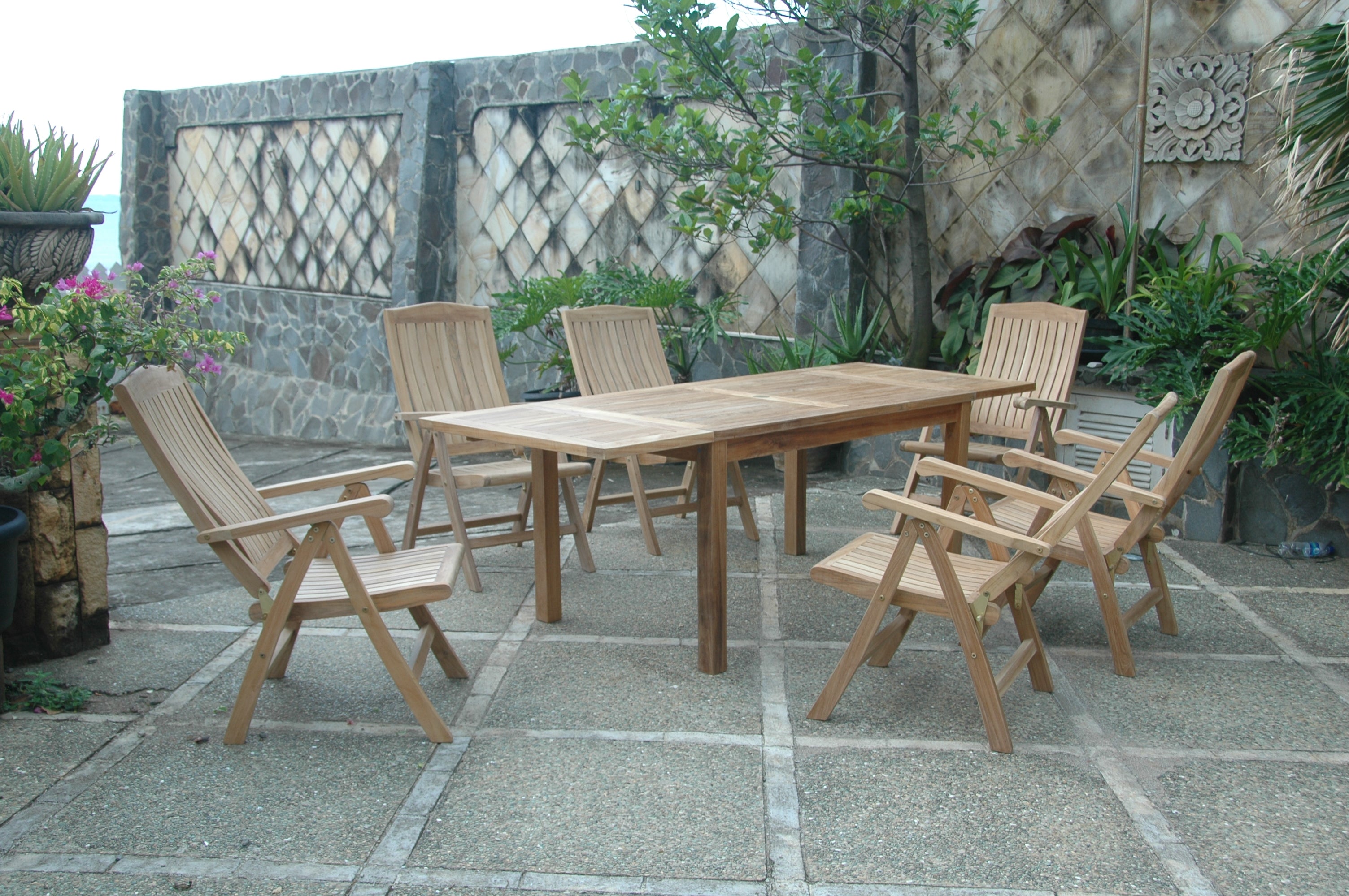 Bahama Katana teak extension table with six reclining armchairs on a stone patio beside a stone garden wall with lattice panels and tropical plants