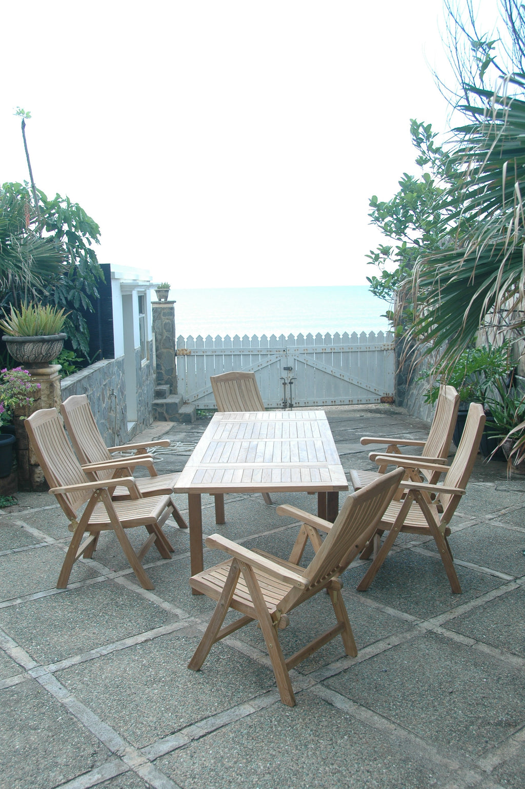 Vertical view of the Bahama Katana teak extension dining set with six reclining armchairs on a patio looking out toward the ocean