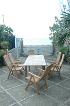 Vertical view of the Bahama Katana teak extension dining set with six reclining armchairs on a patio looking out toward the ocean
