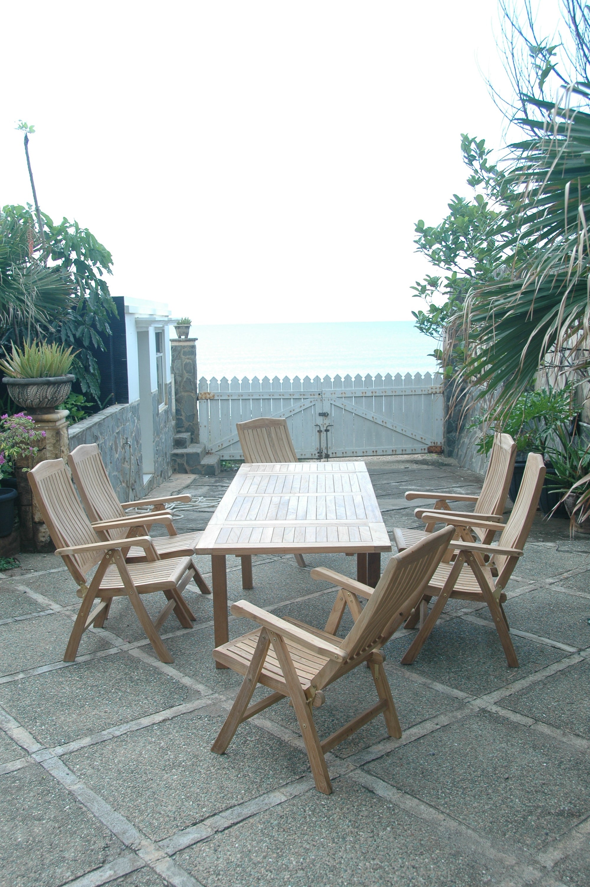 Vertical view of the Bahama Katana teak extension dining set with six reclining armchairs on a patio looking out toward the ocean