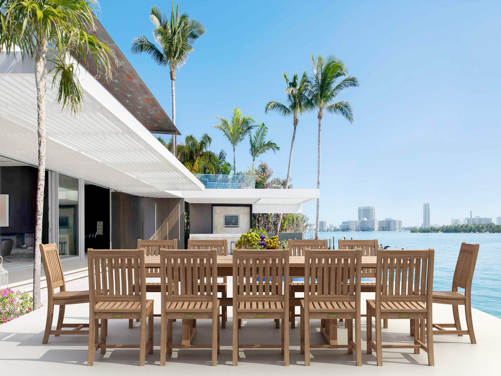 Sahara Rialto teak extension dining table ringed with armless slat-back teak chairs on a white oceanfront terrace, palm trees and a waterfront city skyline beyond
