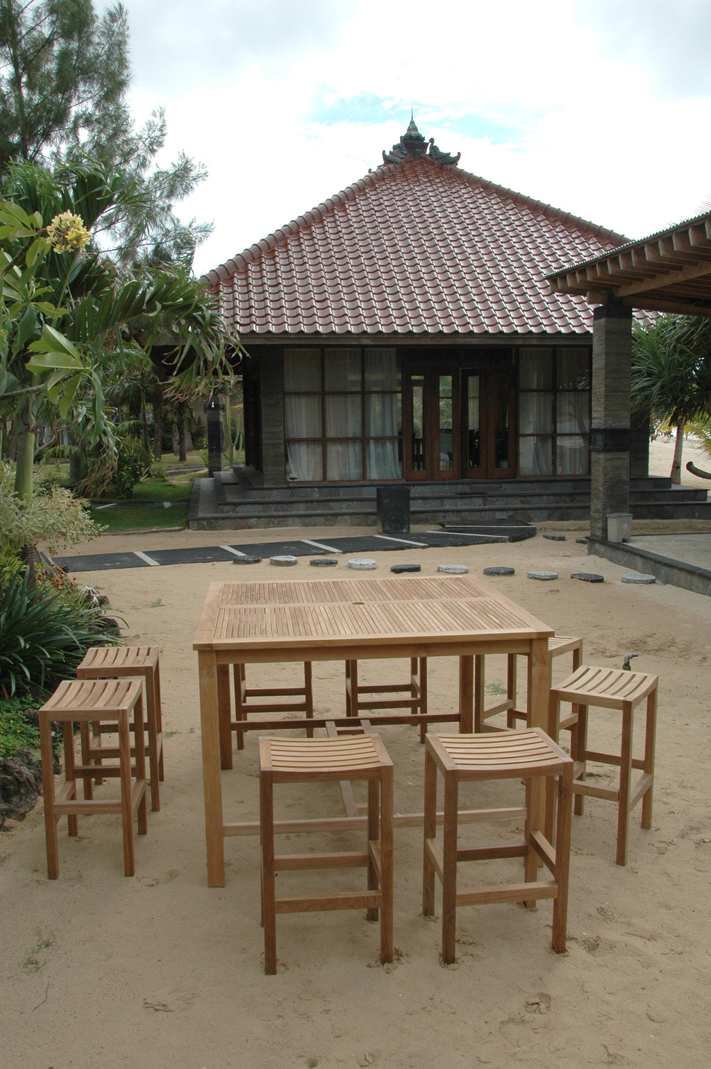 Portrait view of a Windsor square teak bar table with eight Montego backless bar chairs on a garden patio