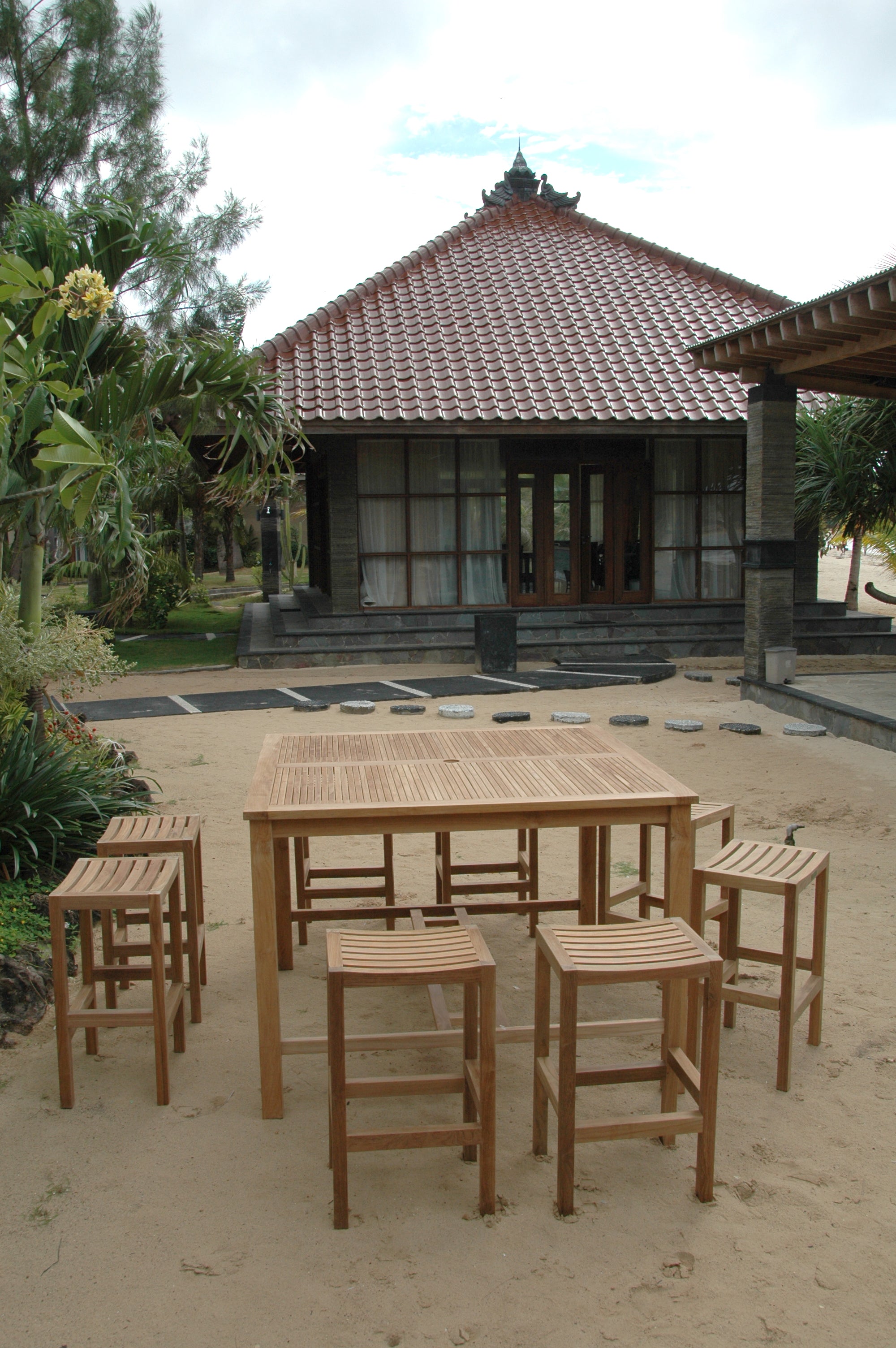 Portrait view of a Windsor square teak bar table with eight Montego backless bar chairs on a garden patio