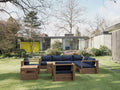 Copacabana modular teak deep seating set arranged as an L-shaped sectional with navy cushions, plus a matching armchair, cushioned ottoman, teak coffee table and side table on a lawn beside a mid-century glass-walled house