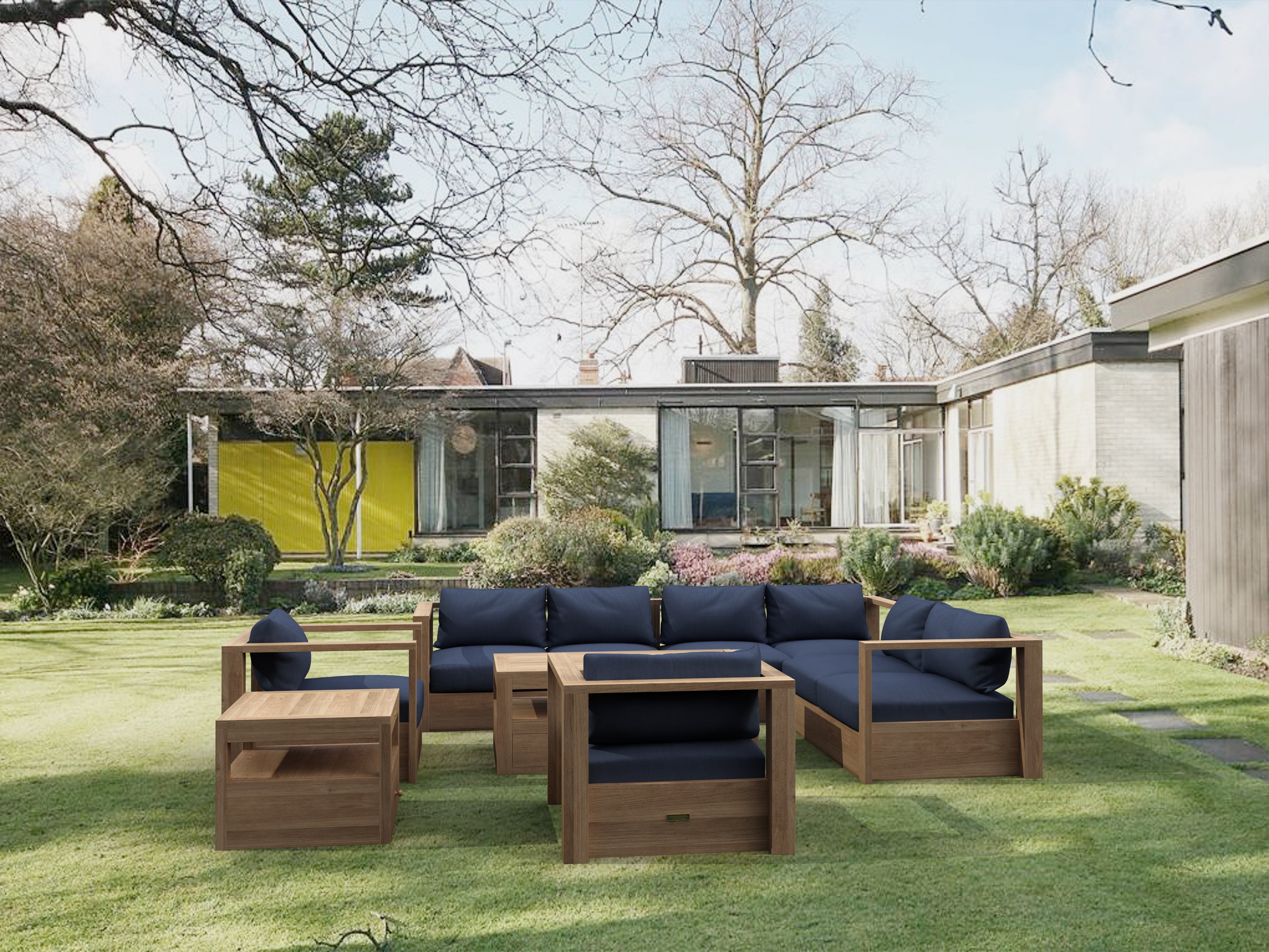 Copacabana modular teak deep seating set arranged as an L-shaped sectional with navy cushions, plus a matching armchair, cushioned ottoman, teak coffee table and side table on a lawn beside a mid-century glass-walled house