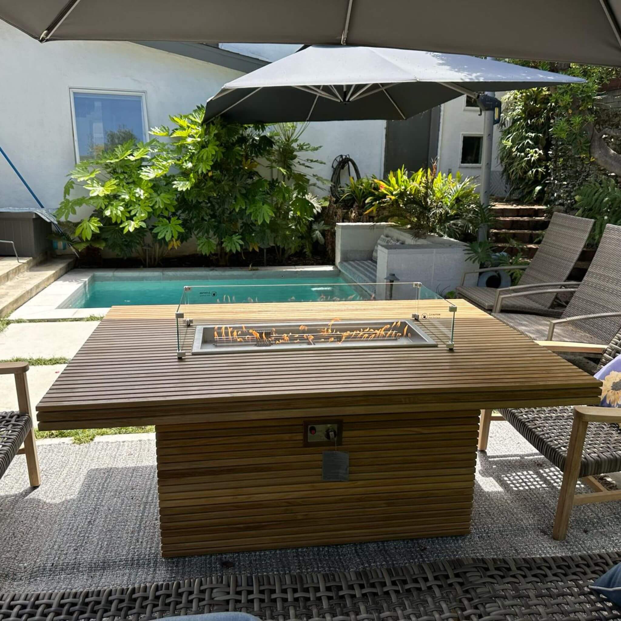 Poolside view of the Louvre teak fire table, showing the lit glass wind guard and controls on the base.
