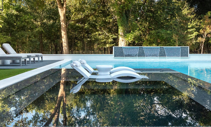Two white in-pool chaise lounges with side tables on a reflective tanning ledge beside a modern pool.